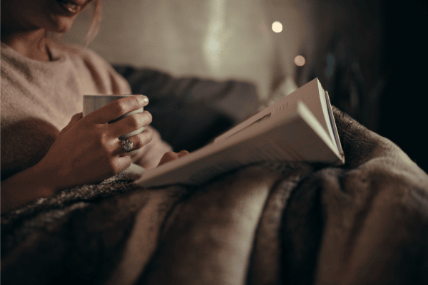 Femme lisant un roman confortablement installée sous un plaid, une tasse à la main